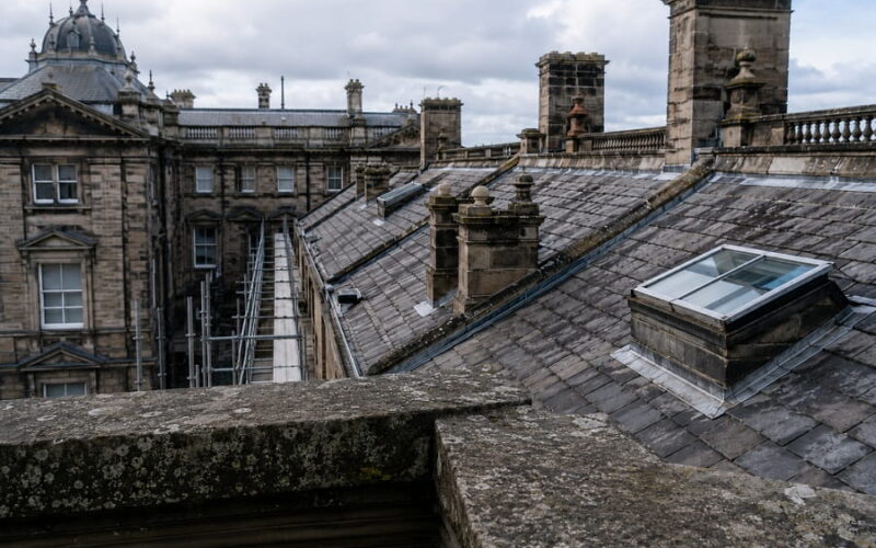 Historic listed commercial building with weathered slate roofs, stone chimneys, rooflights and scaffolding for planned roof repair works.