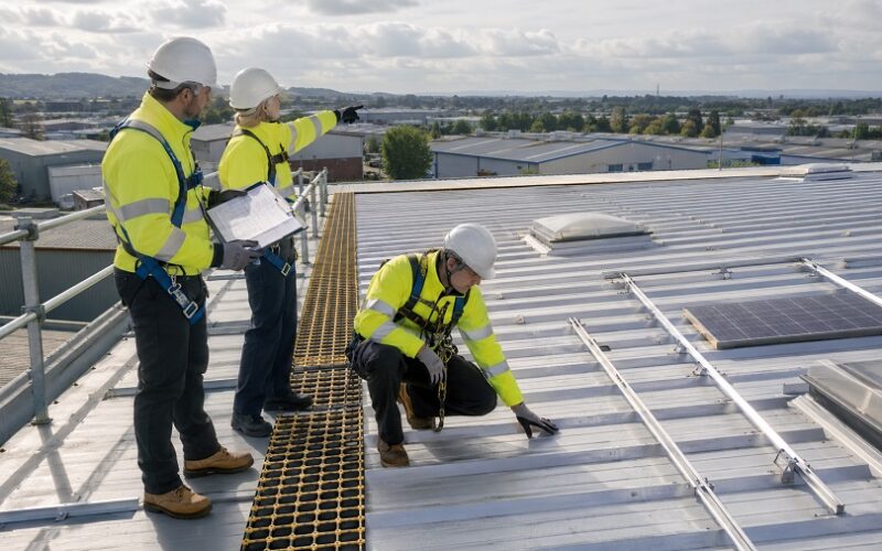 Roofing team in PPE inspecting an industrial roof with rooflights and solar mounting rails during a solar PV roof readiness survey.